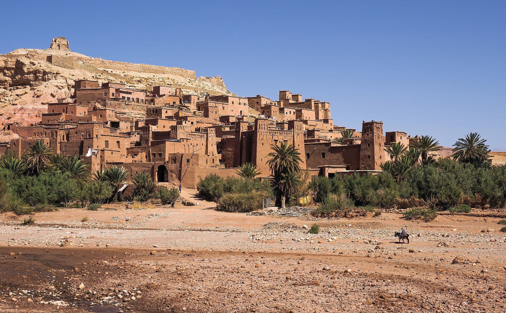 De lemen ksar van Aït Benhaddou tegen een heuvel, met de rivierbedding en palmen op de voorgrond